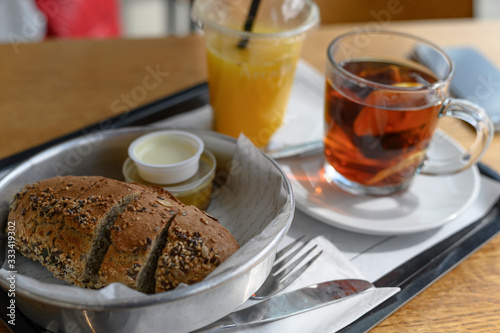 On a tray lies freshly baked sliced bread with seeds, next to butter and sauce. Black tea with lemon and freshly orange juice with a straw in a disposable glass. Breakfast or meal in the cafeteria.