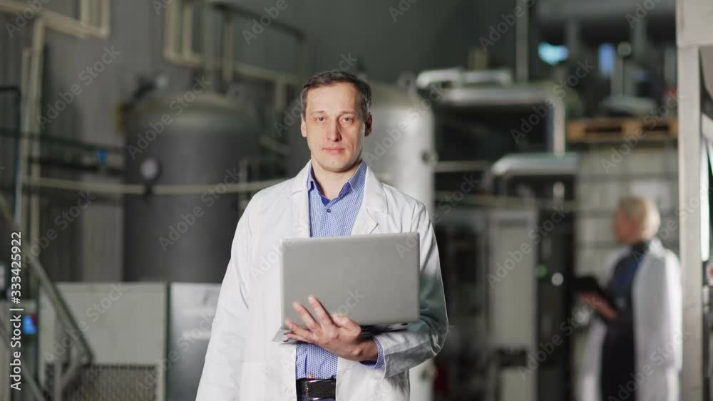 Waist up portrait of middle aged engineer with laptop wearing white coat and looking at camera standing in factory production department Waist up portrait of middle aged engineer with laptop wearing white coat and looking at camera standing in factory production department