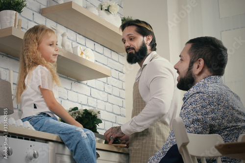 Cheerful same sex male couple with daughter at home. Homosexual male couple cooking in kitchen and talking with their daughter photo