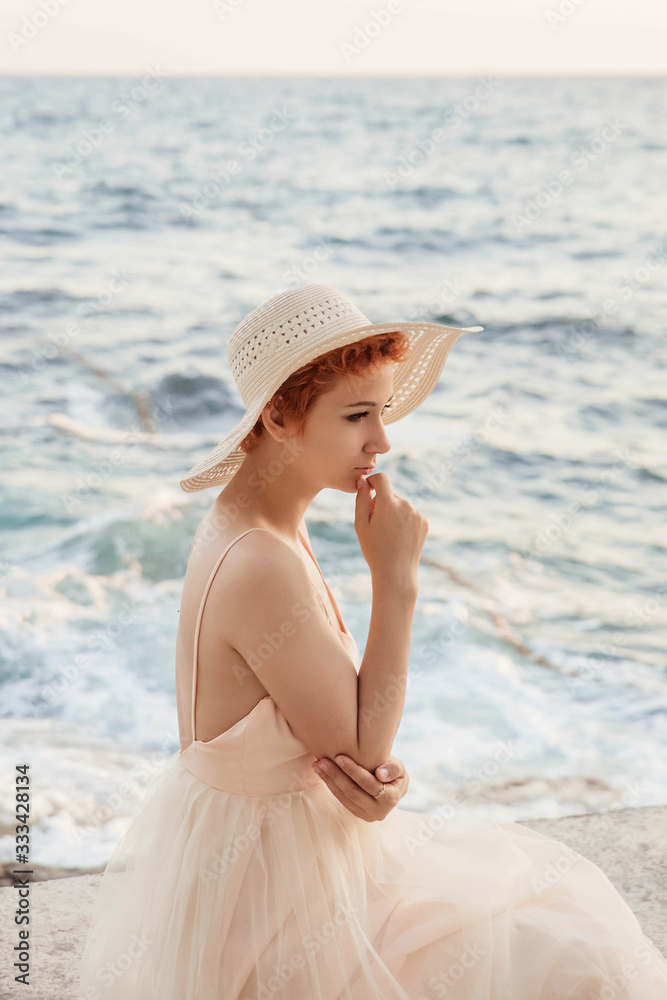 A beautiful, red-haired girl is standing against the sea, wearing a hat on her head, wearing an airy, light dress. Waiting for a lover, the heroine of the novel.