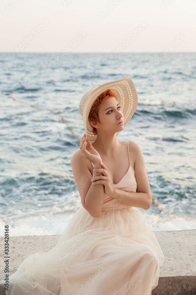 A beautiful, red-haired girl is standing against the sea, wearing a hat on her head, wearing an airy, light dress. Waiting for a lover, the heroine of the novel. A beautiful, red-haired girl is standing against the sea, wearing a hat on her head, wearing an airy, light dress. Waiting for a lover, the heroine of the novel.