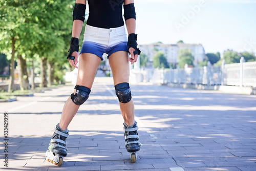 Young woman, roller-blading park with green trees in summer morning at sunny weather. Tan legs, wearing roller-skates and protective equipment, showing the process of riding in town. © Natalia