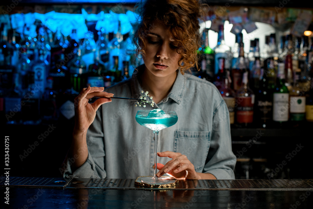 Pretty woman barman decorates glass with blue drink flower branch and look at it.