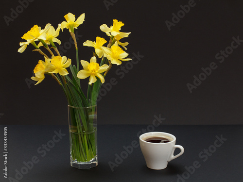 Bouquet of yellow daffodils in vase with cup of coffee on a black background.