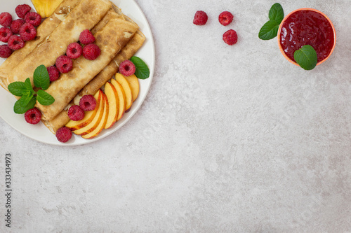Crepes, thin pancakes with raspberries and peaches, mint and jam on a plate. Light background. Top view. Flat lay. Copy space.
