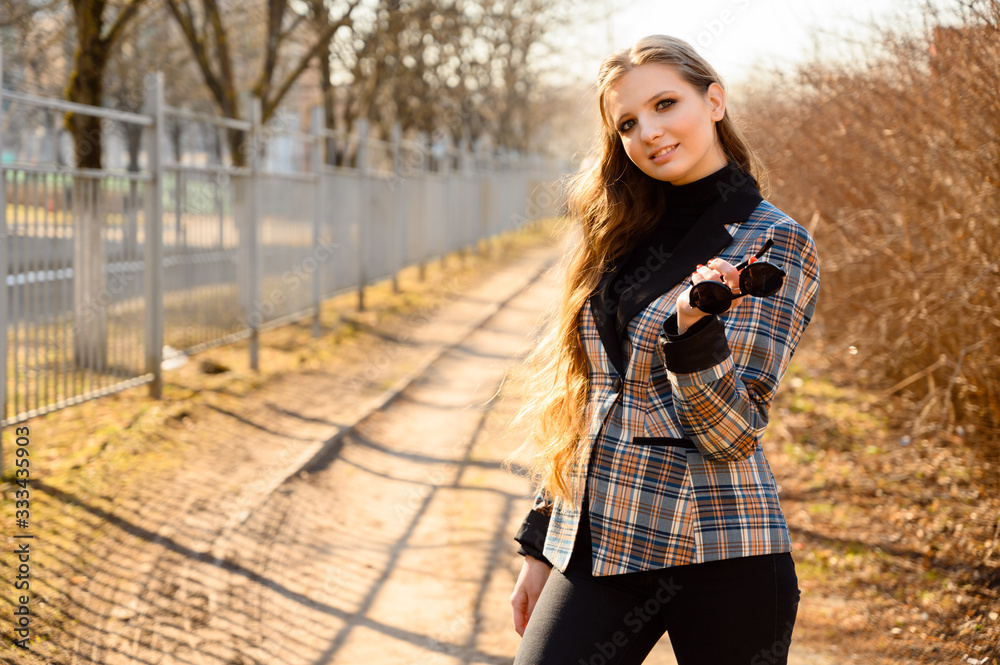 Portrait of a stylish beautiful young girl with long hair with a smile in the park on the background of the road. Photo taken in sunny weather outdoors in spring. Portrait of a stylish beautiful young girl with long hair with a smile in the park on the background of the road. Photo taken in sunny weather outdoors in spring.