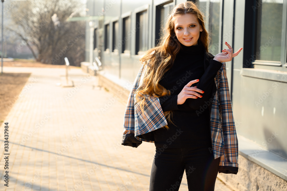 Portrait of a pretty beautiful young girl with long hair with a smile in the city against the background of the wall of a business building. Photo taken in sunny weather outdoors in spring. Portrait of a pretty beautiful young girl with long hair with a smile in the city against the background of the wall of a business building. Photo taken in sunny weather outdoors in spring.