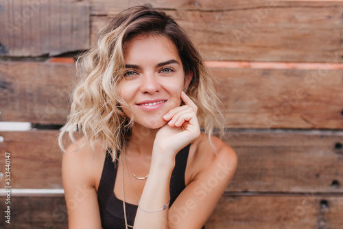 Close-up portrait of dreamy tanned woman with big blue eyes. Photo of lovely young lady touching her face and smiling on wooden background.