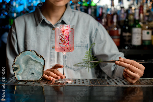 Lady barman begin to decorate glass with rose cocktail by piece of green plant.