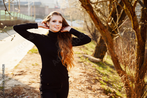 Portrait of a stylish young woman with long hair with a smile on the background of road trees in the park. Photo in sunny weather outdoors in spring.