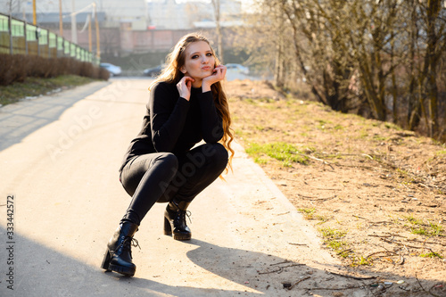 Portrait of a pretty girl crouched over the high road of a young girl with long hair with a smile on a city background. Photo in sunny weather outdoors in spring. © Вячеслав Чичаев