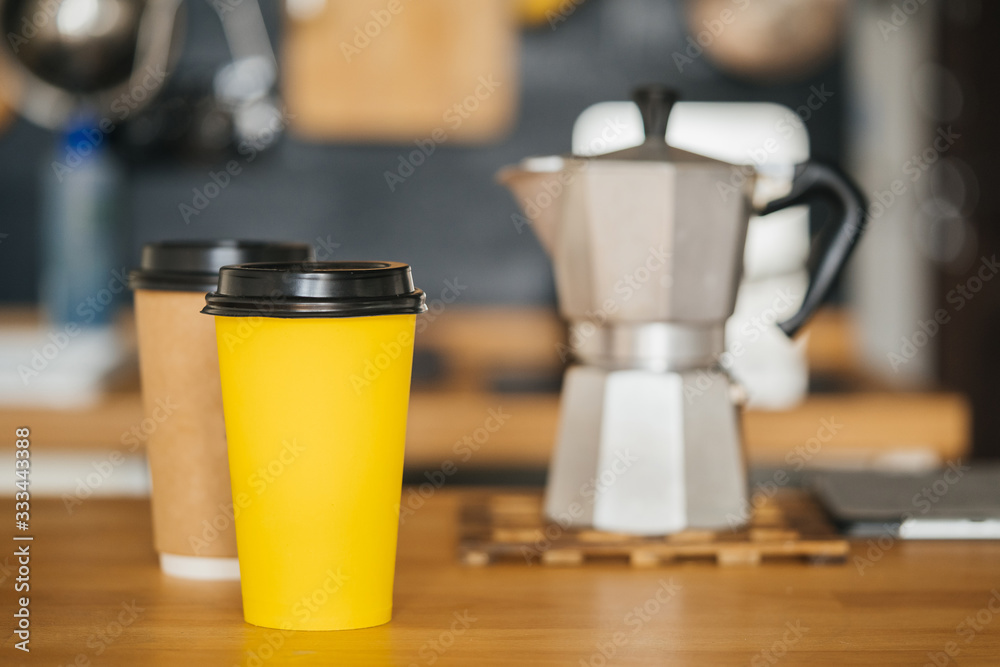 A paper yellow glass under coffee is on the table of a modern loft kitchen against the background of a geyser coffee maker. Takeaway morning coffee.