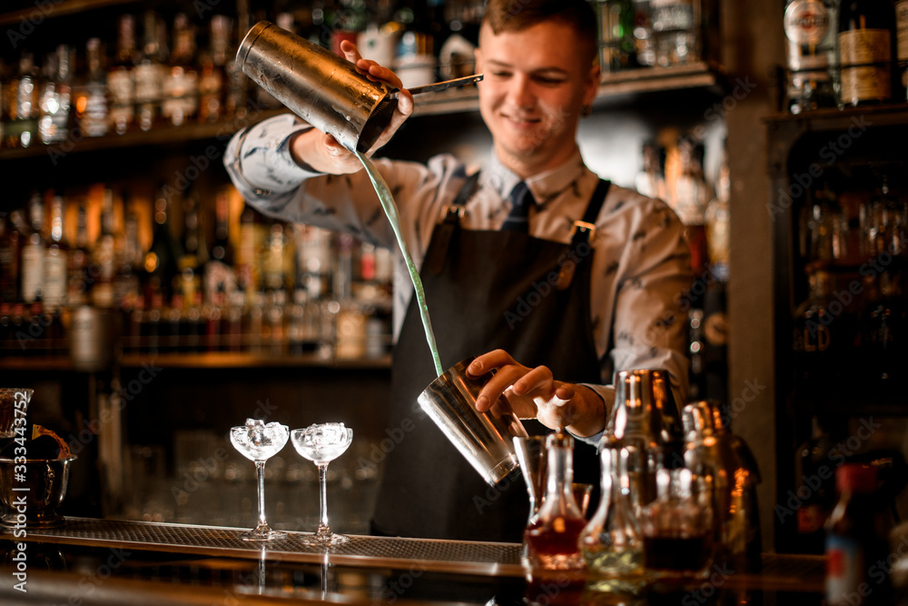 Young smiling barman pours cocktail in shaker.