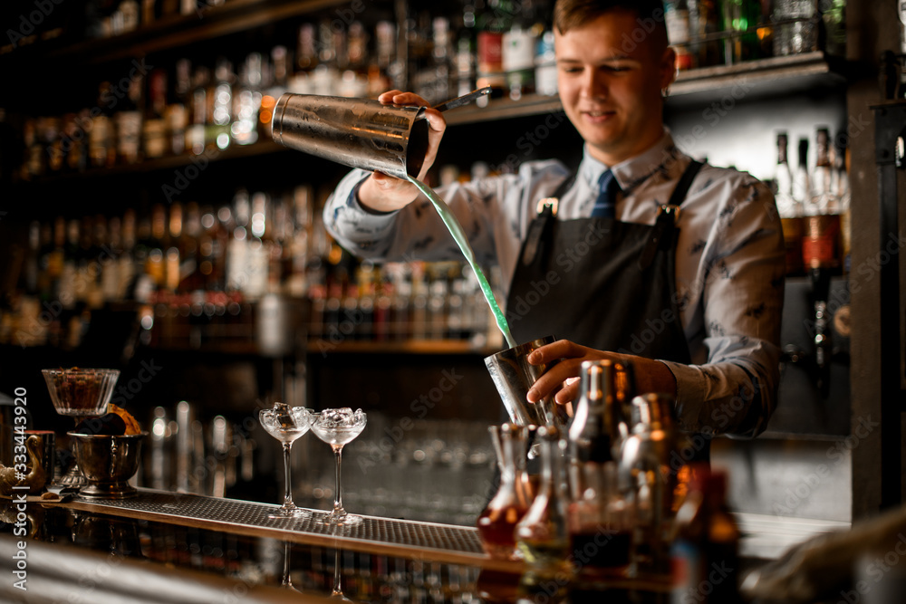 Young smiling barman pours cocktail in steel shaker.