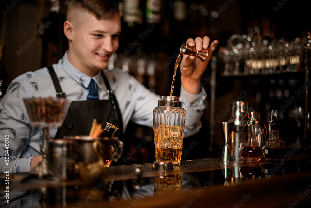 Young smiling bartender carefully pours drink to glassy shaker using beaker.