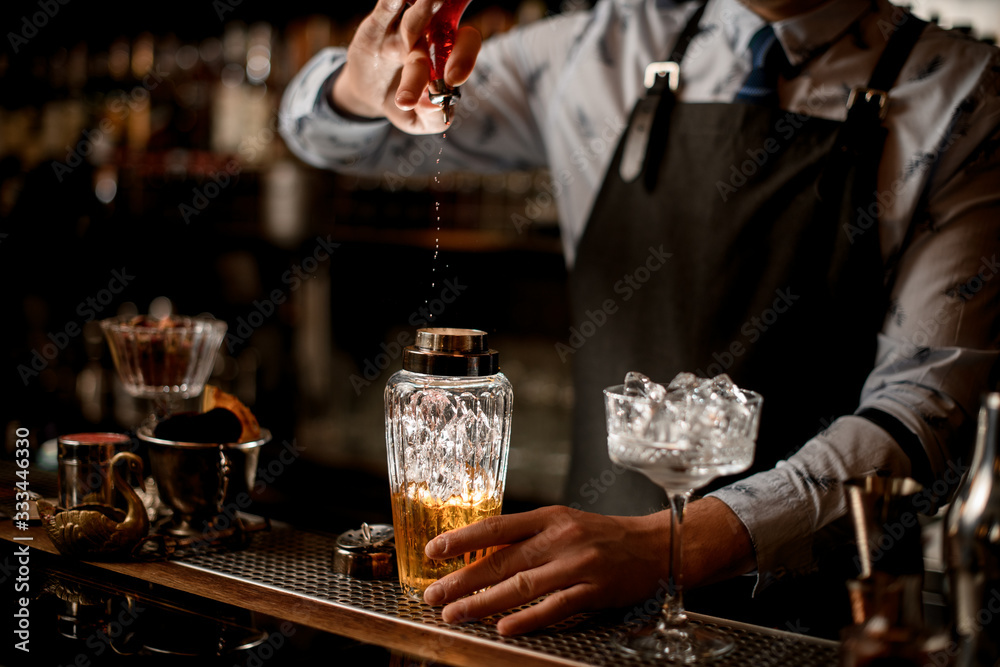 Bartender in black apron actively pours drink to glassy shaker.