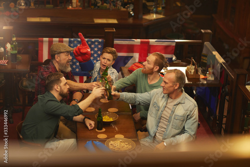 Group of men toasting with glasses of beer while woman cheering for the sport team they sitting at the table in sport bar