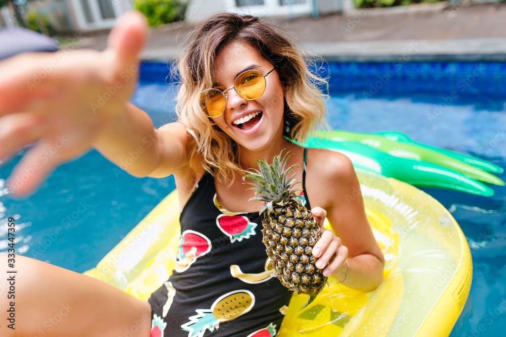 Amazing short-haired girl in trendy glasses swimming in pool. Outdoor photo of lovely tanned female model in stylish swimsuit laughing in summer weekend. - obrazy, fototapety, plakaty 