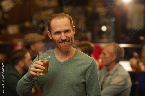 Portrait of young bearded man drinking beer and smiling at camera while standing in the pub