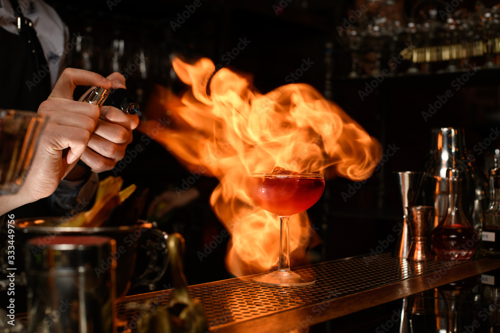 Bartender professionally sprinkles on glass with cocktail and sets it on fire.
