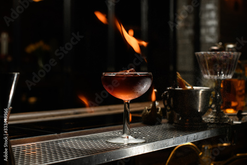 glass with brown alcoholic drink and piece of ice stands on the bar. Flame above it.