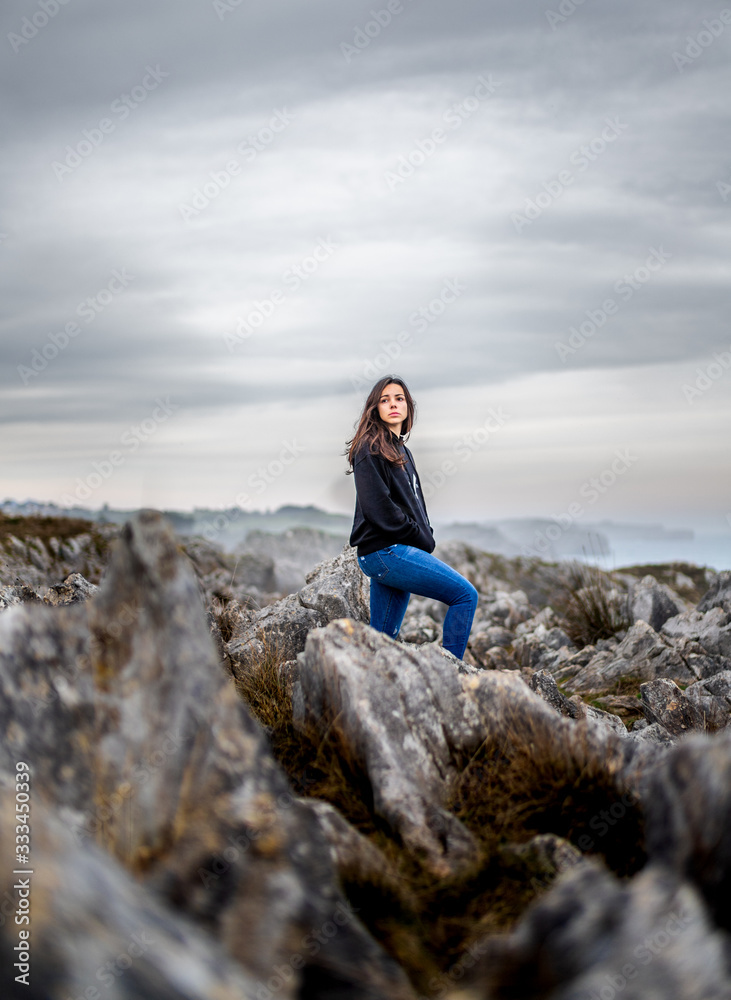 Beautiful young girl with black sweater standing on rocks beside cliffs at the coast in a beautiful landscape Beautiful young girl with black sweater standing on rocks beside cliffs at the coast in a beautiful landscape