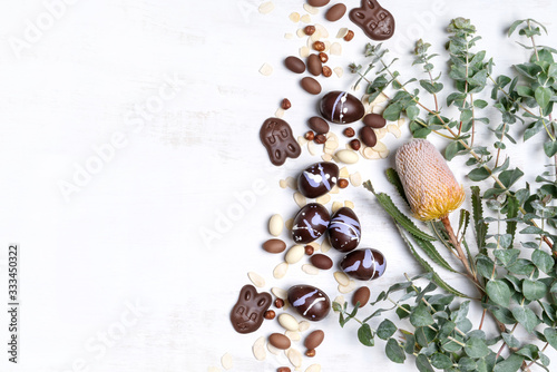 Fototapeta Naklejka Na Ścianę i Meble -  Vegan chocolate Easter eggs and Easter bunnies surrounded by shaved almonds, hazelnuts plus Australian native Banksia and Eucalyptus leaves on a rustic white background.