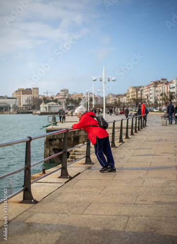 Old woman leaning on a fence looking to the sea in a small town of the north of spain in a sunny day in winter © Alex