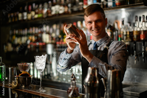 Young smiling bartender standing behind the bar counter with steel shaker.