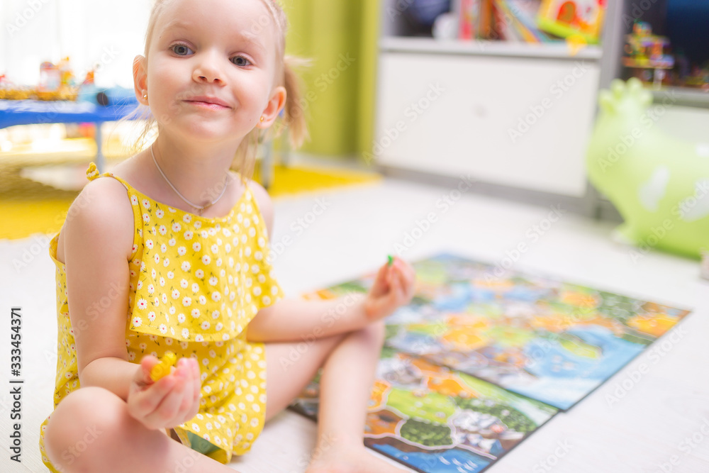 Fototapeta premium Board game concept. Little kid girl play in board game at home sitting on a wooden floor. Board game field, many figures.