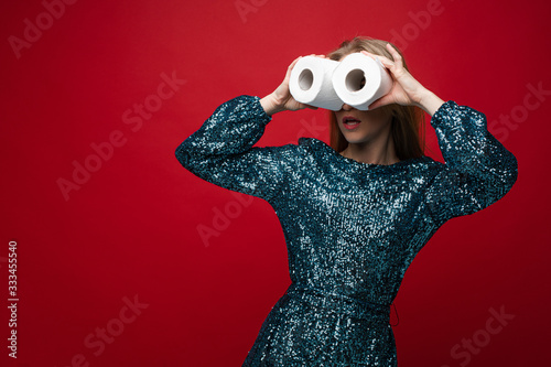 Fashion woman in shining dress posing looking through toilet paper rolls isolated at red studio background. Glamour female holding hygiene accessories like binoculars. Coronavirus infection concept
