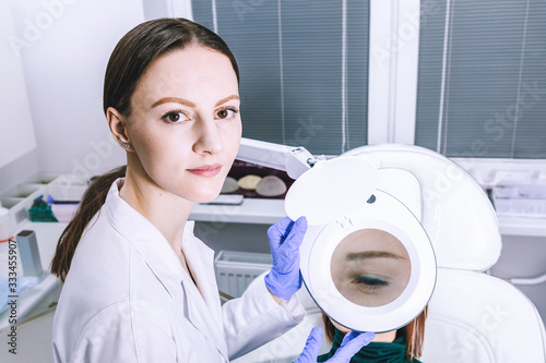 Female woman doctor is analyzing skin through magnifier on female patient face before the procedure in cabinet. Beautician cosmetology concept, presentation of anti-aging skin treatment photo