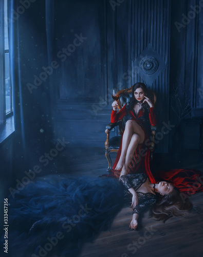 Art evil woman vamp. Red long dress sits in vintage armchair. Holding glass Blood wine. at feet lies sleeping beauty dead princess. black lush dress. Bloody vampire bite. backdrop medieval room night