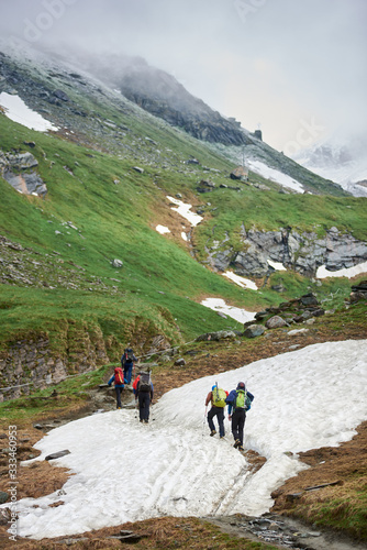 Back view of travelers with backpacks and trekking sticks walking on mountain road. Group of active people climbing snowy hill one after another. Concept of traveling, hiking and mountaineering.