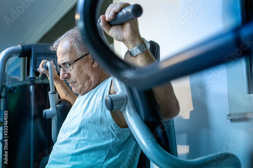 Uomo anziano con canotta celeste si allena in palestra ai macchinari photo