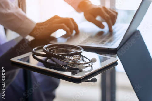 Male doctor hands typing on laptop computer keyboard, search medical information with digital tablet pc and medical stethoscope on the desk at office. Online medical,medic tech, emr, ehr concept.   photo