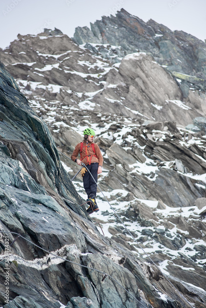 Full length of male alpinist in sunglasses and safety helmet ascending high mountain. Man climber holding fixed rope and looking down. Concept of mountaineering, alpinism and climbing.