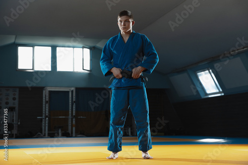 Young judo caucasian fighter in blue kimono with black belt posing confident in the gym, strong and healthy. Practicing martial arts fighting skills. Overcoming, reaching target, self building up. photo