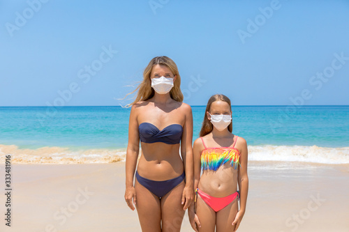 A woman and a child in a protective surgical mask on their face are standing on the beach in swimsuits. Chinese coronavirus disease COVID-19 is dangerous © dadoodas