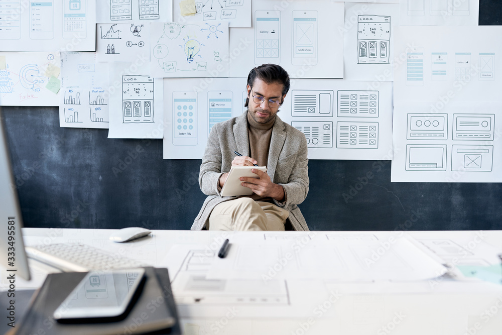Busy fashionable man in glasses sitting against board with interface sketches and working on new design project Busy fashionable man in glasses sitting against board with interface sketches and working on new design project