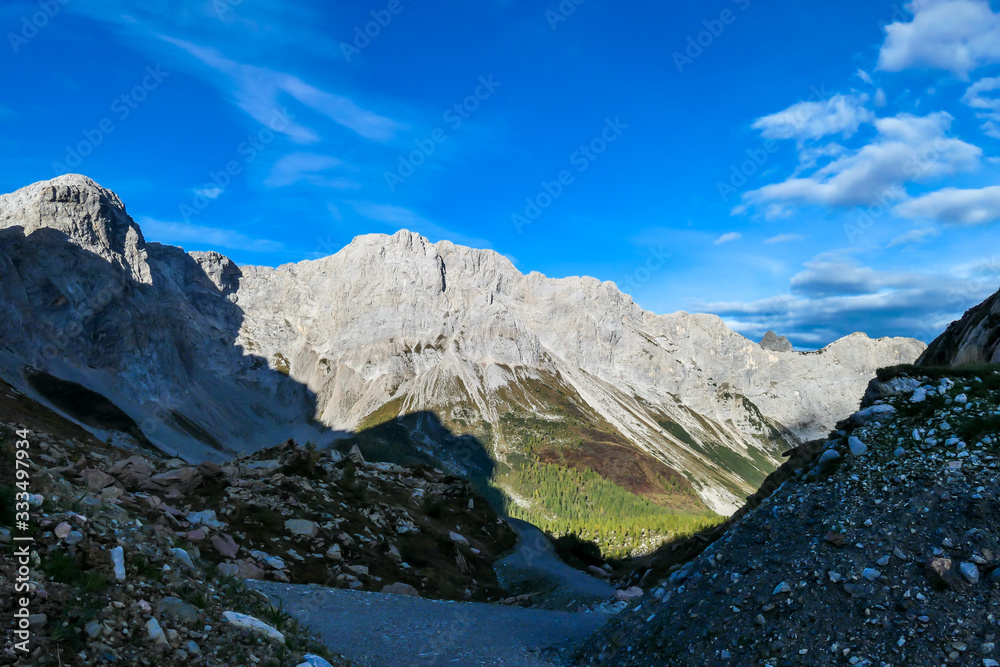 Obraz premium A pathway leading to high peaks in Italian Alps. Sharp slopes on both sides of the valley. Hard to reach mountain peaks. There are many mountain ranges in the back. Serenity and peace. Autumn vibes