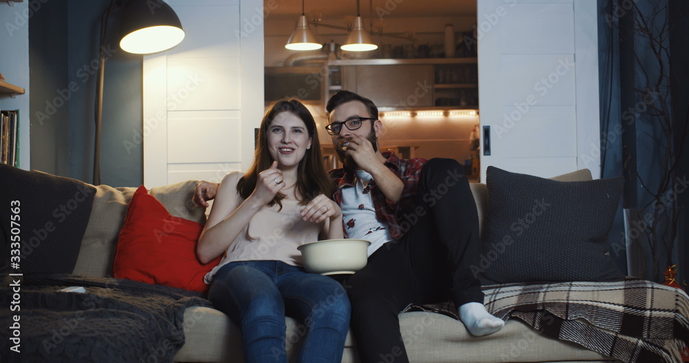 Happy young casual European romantic couple watching TV series at home, smiling and talking, eating popcorn slow motion Happy young casual European romantic couple watching TV series at home, smiling and talking, eating popcorn slow motion