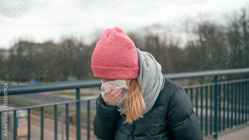 COVID-19 Coronavirus Pandemic Outbreak: Casual Young Sick Woman in Surgical Face Mask and Medical Gloves Sneezes at Street and Coughs covering mouth