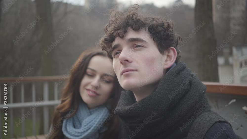 Close-up of young curly-haired Caucasian man looking up and smiling as girlfriend putting head on his shoulder at the background. Happiness, lifestyle, love, romance. Close-up of young curly-haired Caucasian man looking up and smiling as girlfriend putting head on his shoulder at the background. Happiness, lifestyle, love, romance.