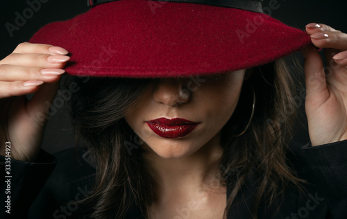 Close-up of sensual glamorous beauty hiding face under hat touching maroon wide brim with both hands photo