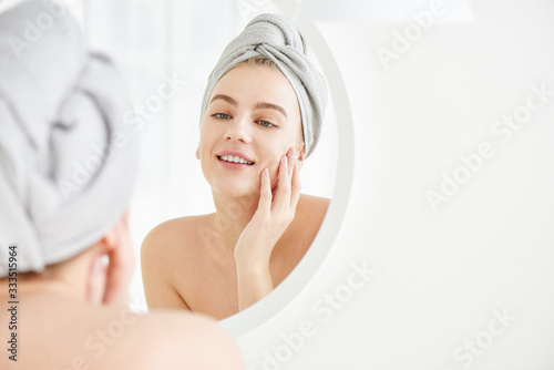 Portrait of  young girl with  towel on head in white bathroom looks and touches her face in the mirror and enjoys youth and hydration. Natural beauty, home care for problem skin photo