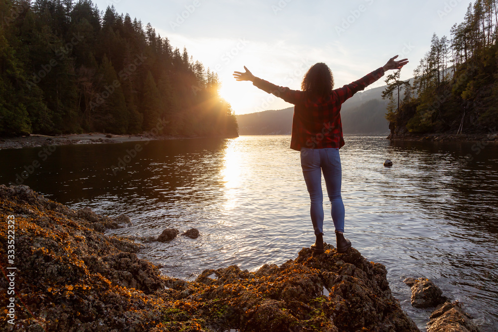 Fototapeta premium Adventurous Girl with Hands Up in the Canadian Landscape during a vibrant winter sunset. Hike on Jug Island Trail in Belcarra, Vancouver, British Columbia, Canada. Concept: Freedom, Explore, Health