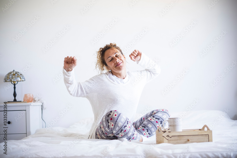 Early morning wake up concept with happy cheerful caucasian beautiful adult woman stretching and smiling - white colors and light in the bedroom athome or hotel - stay home quarantine - obrazy, fototapety, plakaty 