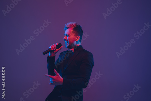 Young caucasian musician, performer singing on gradient purple-blue background in neon light. Concept of music, hobby, festival. Joyful party host, DJ, stand upper, singer. Colorful portrait of artist