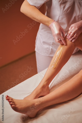 Waxing. The specialist removes hair from the beautiful legs of a young girl using wax. The girl lies on the sofa in the spa and she is doing depilation.